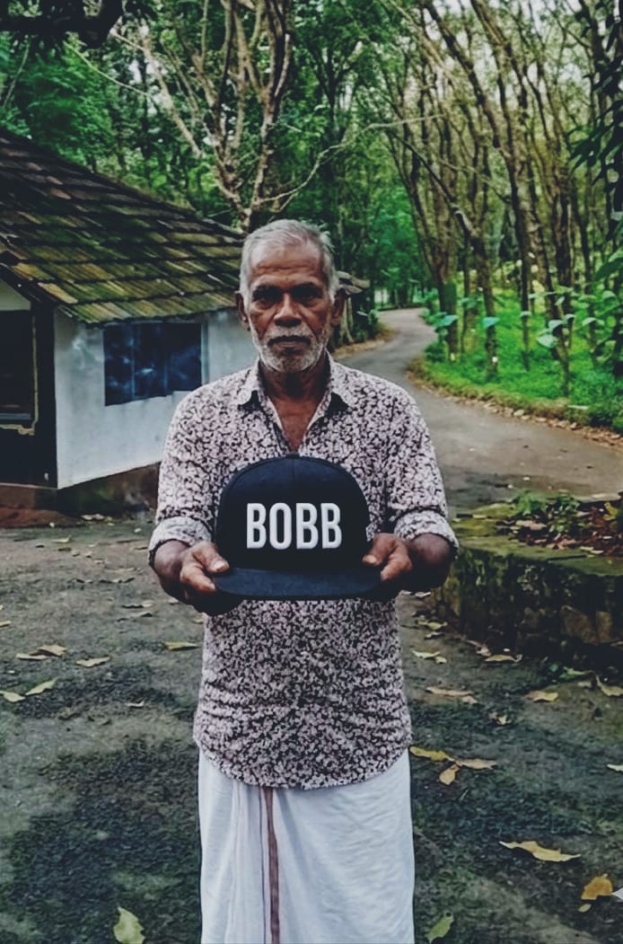A man on a tree-lined Kannur lane holding out a black BOBB cap with both hands